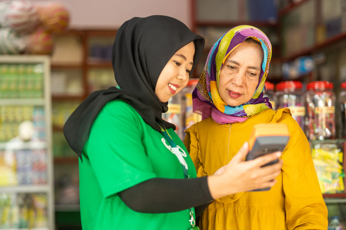 Dua perempuan berdiri di dalam sebuah toko kecil sambil melihat layar ponsel bersama-sama