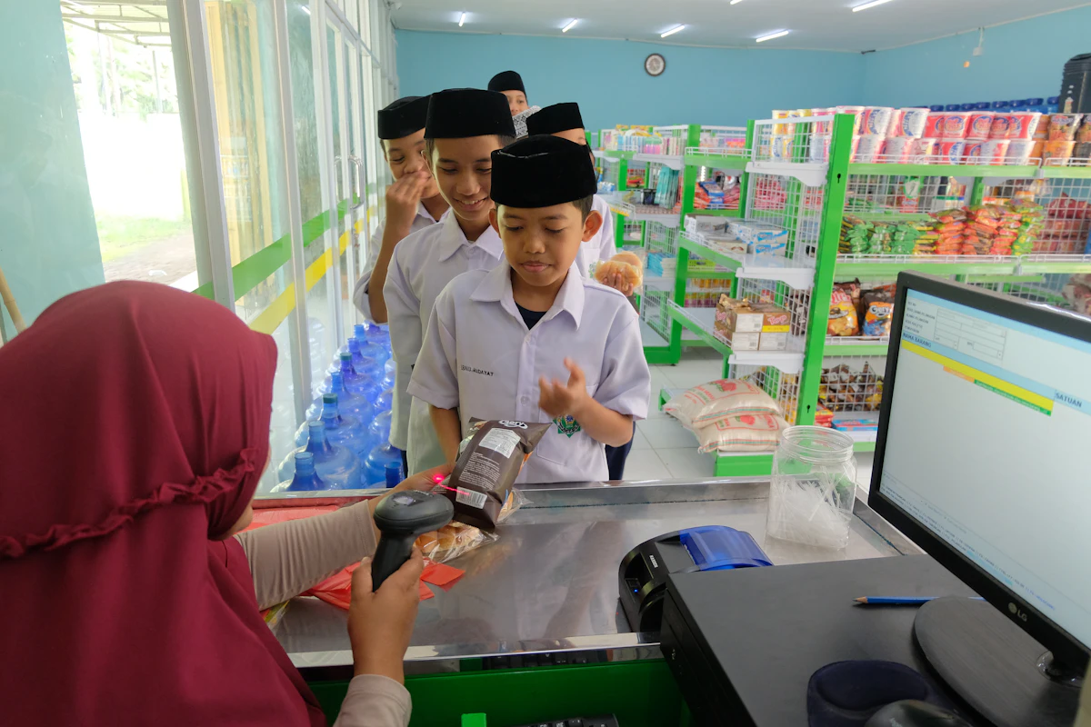 Penjual menyerahkan barang kepada seorang anak di meja depan toko kelontong kecil, rak dagangan penuh di belakangnya