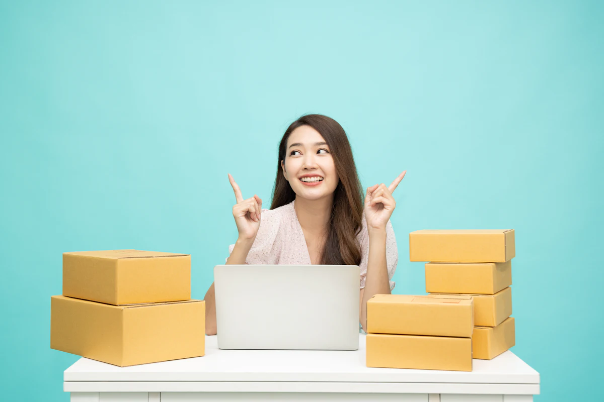 Seorang perempuan duduk di meja dengan laptop terbuka, dikelilingi tumpukan kardus paket