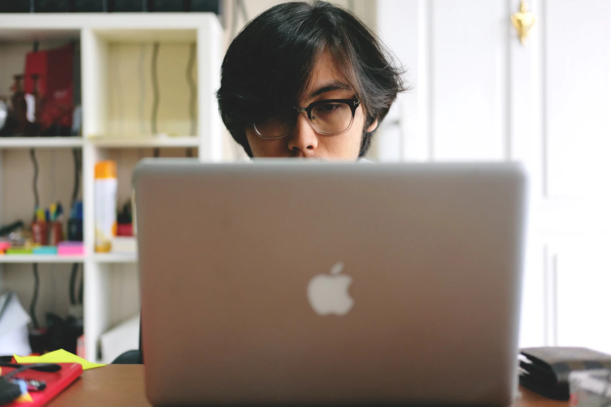 Pria berkacamata bekerja di laptop di sebuah ruangan dengan rak kayu berisi barang di belakangnya