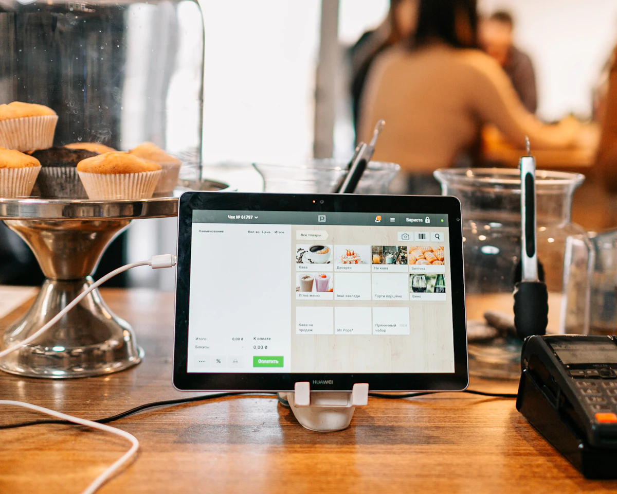 Tablet aplikasi kasir menampilkan daftar produk di atas meja kayu toko, dengan pembaca kartu di sampingnya