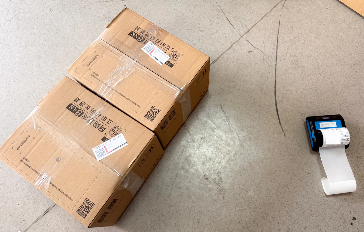 Tumpukan kardus paket siap kirim di meja kerja toko online, dengan printer label kecil di sampingnya, cahaya siang alami, tanpa wajah orang