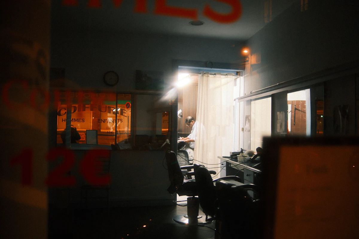 Bagian dalam sebuah barbershop terlihat dari balik kaca jendela pada malam hari, ruangan sudah kosong dan lampunya tinggal sebagian