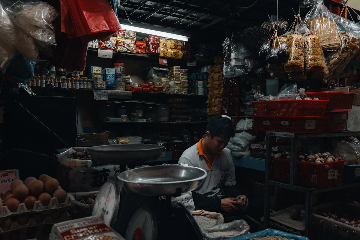 Pemilik toko berdiri di antara rak yang penuh barang dagangan di toko kecilnya